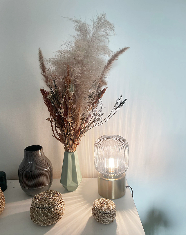 Une lumière douce éclaire des décorations posées sur une table. On y voit un pot de fleurs séchées, ainsi qu'un case et des petits bols tressées en osier.