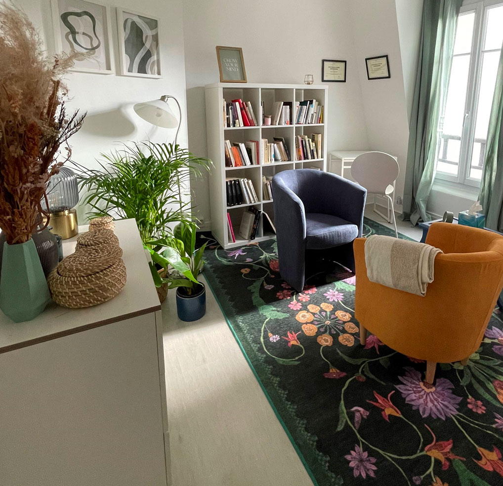 Le cabinet de consultation, on y voit un fauteuil confortable devant une petite bibliothèque, un tapis floral et des plantes égayent la pièce